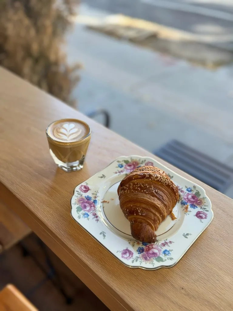 A croissant on a vintage floral plate beside a cortado on a sunlit wooden ledge